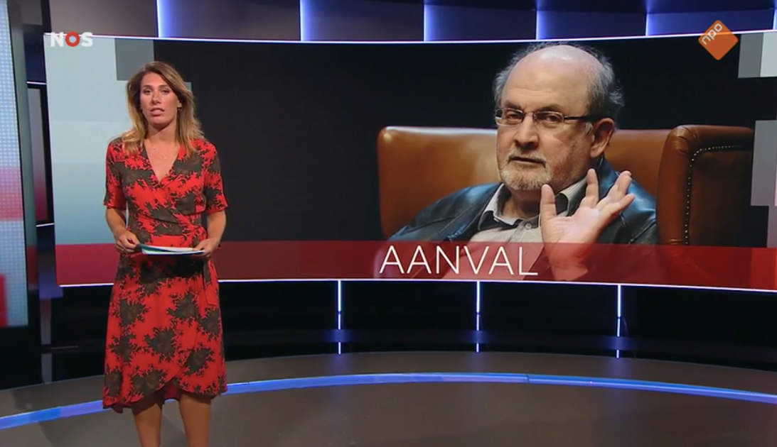 Beeld uit het NOS Journaal toont de nieuwslezer op de voorgrond, met op de achtergrond een portret van Rushdie, met daarbij de tekst &#39; AANVAL&#39; .